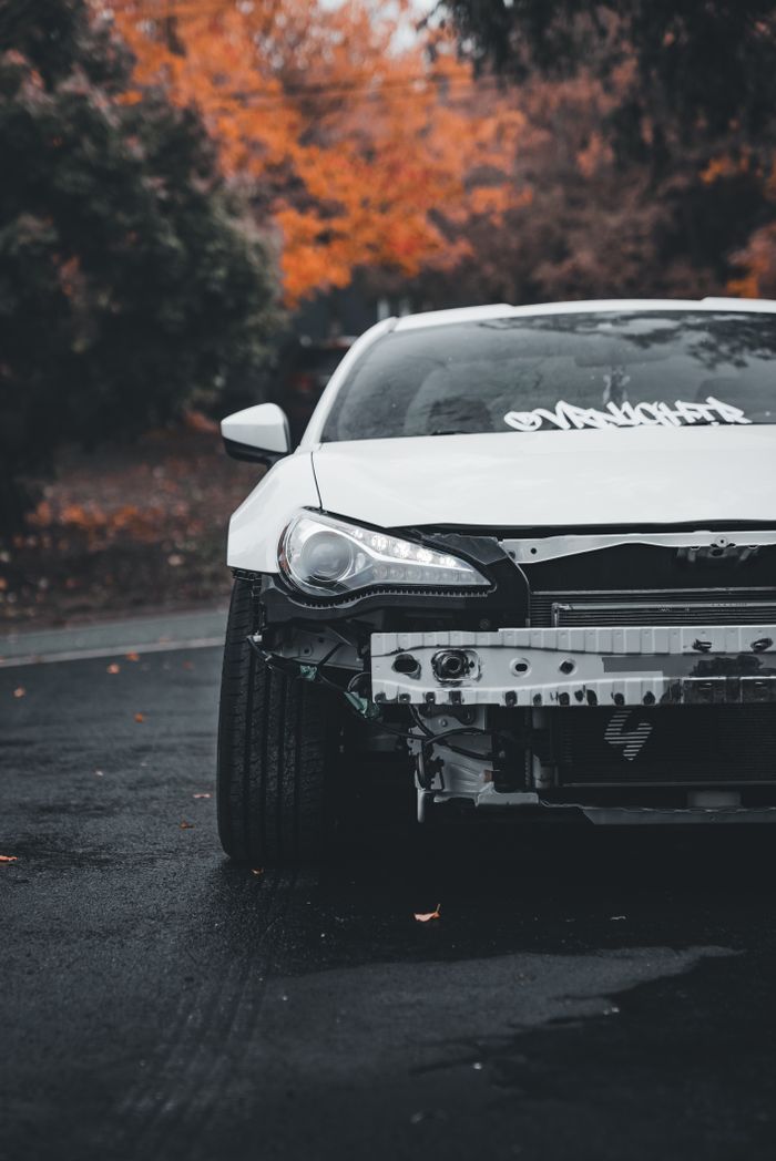 Close-up of a damaged white car on a wet road with autumn foliage in the background.